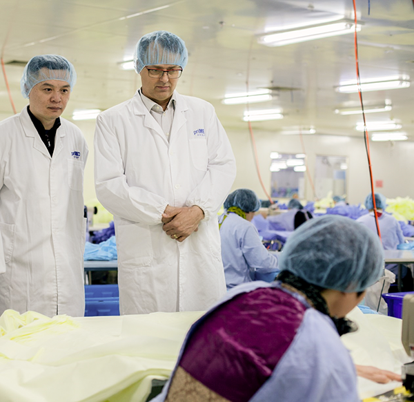 Two people are wearing lab coats and hair nets. They are standing and watching another person wearing a hair net and protective gown work on the production of face masks.