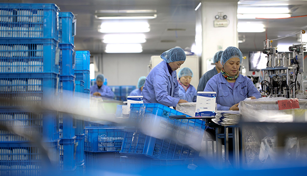 Six people are dressed in gowns and hair nets and working on the production of personal protective equipment. In the foreground are stacks of plastic boxes.