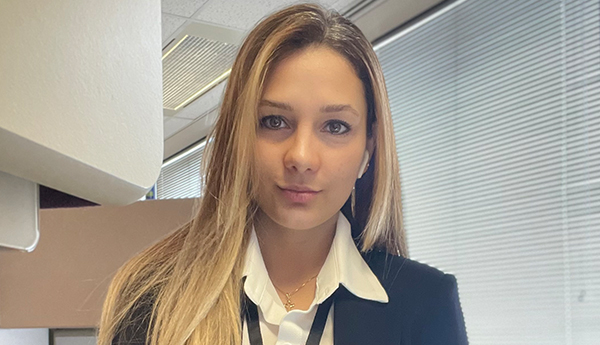 Une jeune femme vêtue d'une chemise blanche et d'un veston marine dans un bureau. Des fauteuils violets sont visibles à l'arrière-plan.