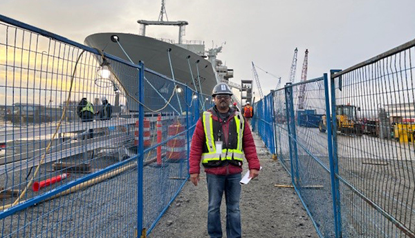 Un homme vêtu d'un gilet de sécurité et d'un casque de protection est debout entre des barrières de sécurité. Un navire militaire est visible à l'arrière-plan.