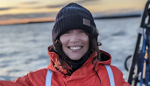 A smiling person on a boat, with water in the background, wearing an orange coat and a wool hat.