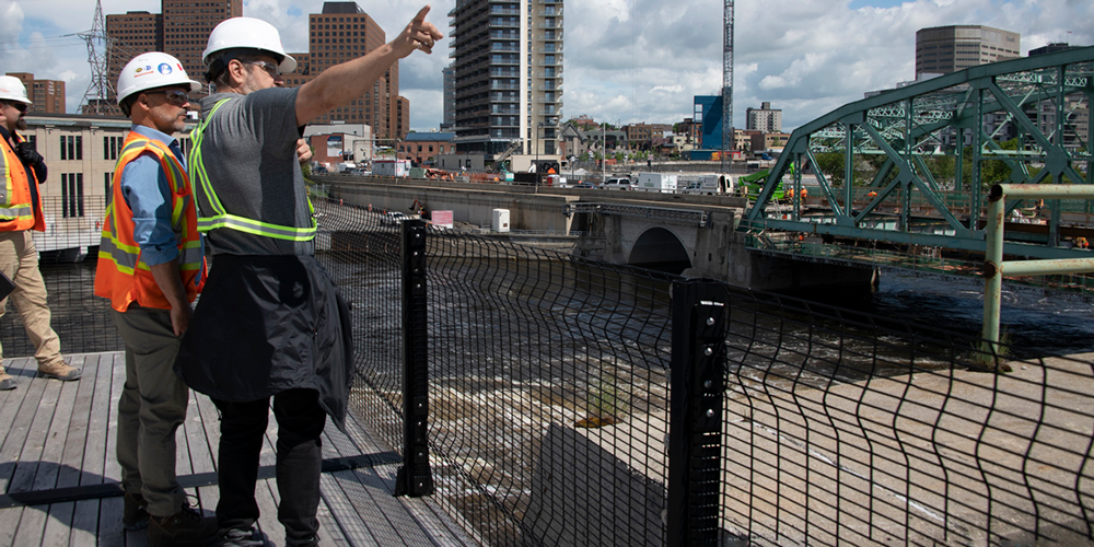 2 people talking on a platform next to a river and one of them is pointing toward the sky.