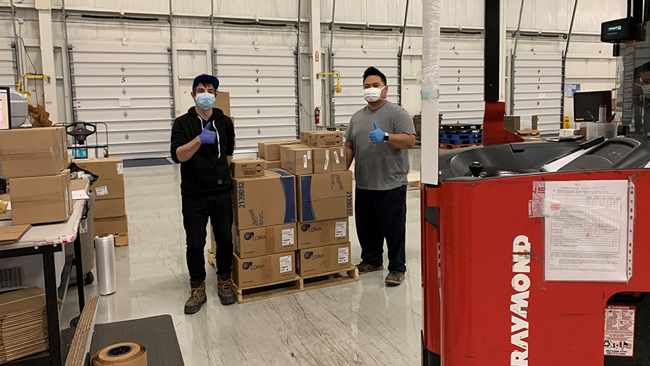 Two people are in a warehouse standing either side of a pile of cardboard boxes. They are wearing masks and gloves and are giving thumbs-up signs.