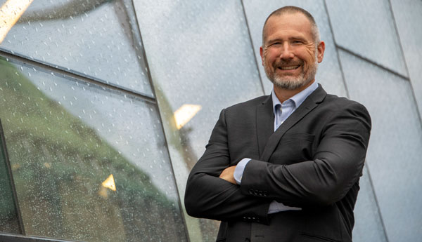A man dressed in a dark suit is standing in front of a glass building with his arms crossed.