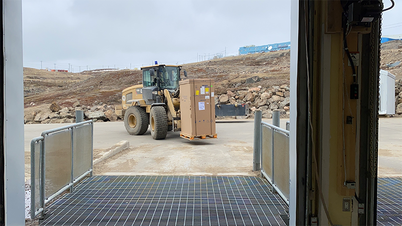 Un chargeur élévateur frontal transporte un grand colis rectangulaire et avance vers une aire de chargement.