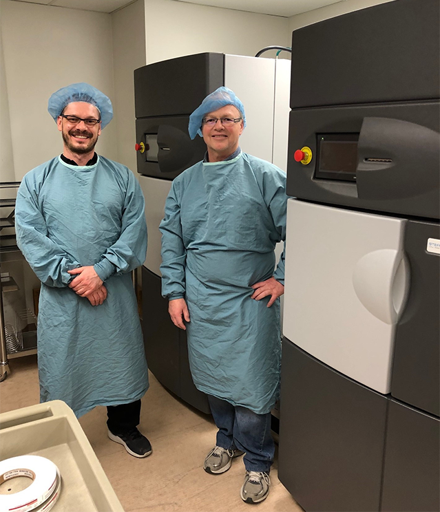 Deux personnes, portant des blouses médicales et des bonnets, sont debout entre deux machines et sourient pour l’appareil photo.