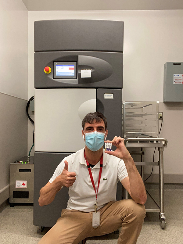 Une personne portant un masque est accroupie et se trouve devant une machine. La personne tient 4 petites fioles et a le pouce de la main droite levé en l’air.