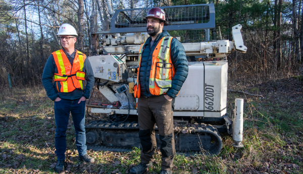 2 men in safety vests and hard hats standing in front of a machine in a grassy area.