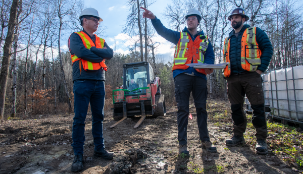 3 men in safety vests and hard hats standing in a dirt clearing. One 1 man is pointing and holding a map.