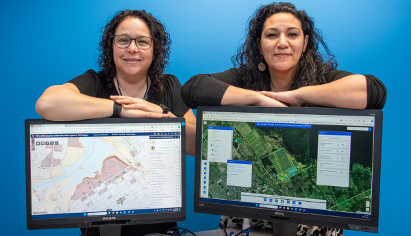2 women against a plain blue background and leaning on computer screens showing maps.