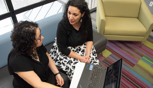 2 women sitting on a bench and having a conversation in front of a computer.