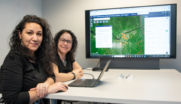 2 smiling women sitting at a desk in front of a computer and a large monitor showing a map.