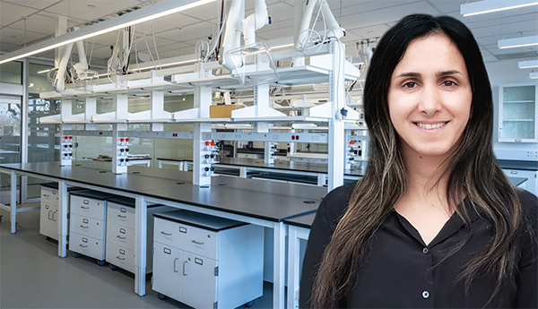 Une femme en blouse noire qui sourit pour une photo. Derrière elle, des tables de laboratoire en métal avec des armoires blanches et une rampe aérienne avec de l'équipement blanc.