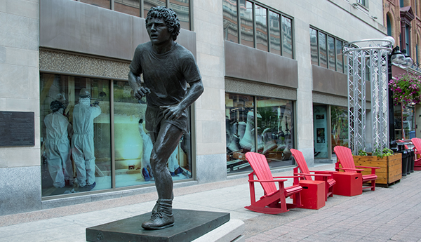 A front view of the Terry Fox bronze statue on a concrete slab, with 3 red Adirondack chairs and a glazed building.