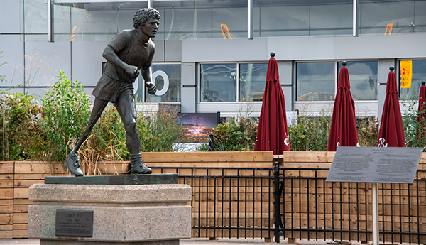 The Terry Fox bronze statue on a concrete slab in front of 3 red sun umbrellas, a wooden fence and a white building.
