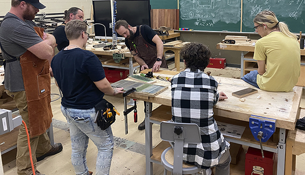 Students listen to their instructor while sitting and standing around a workbench.