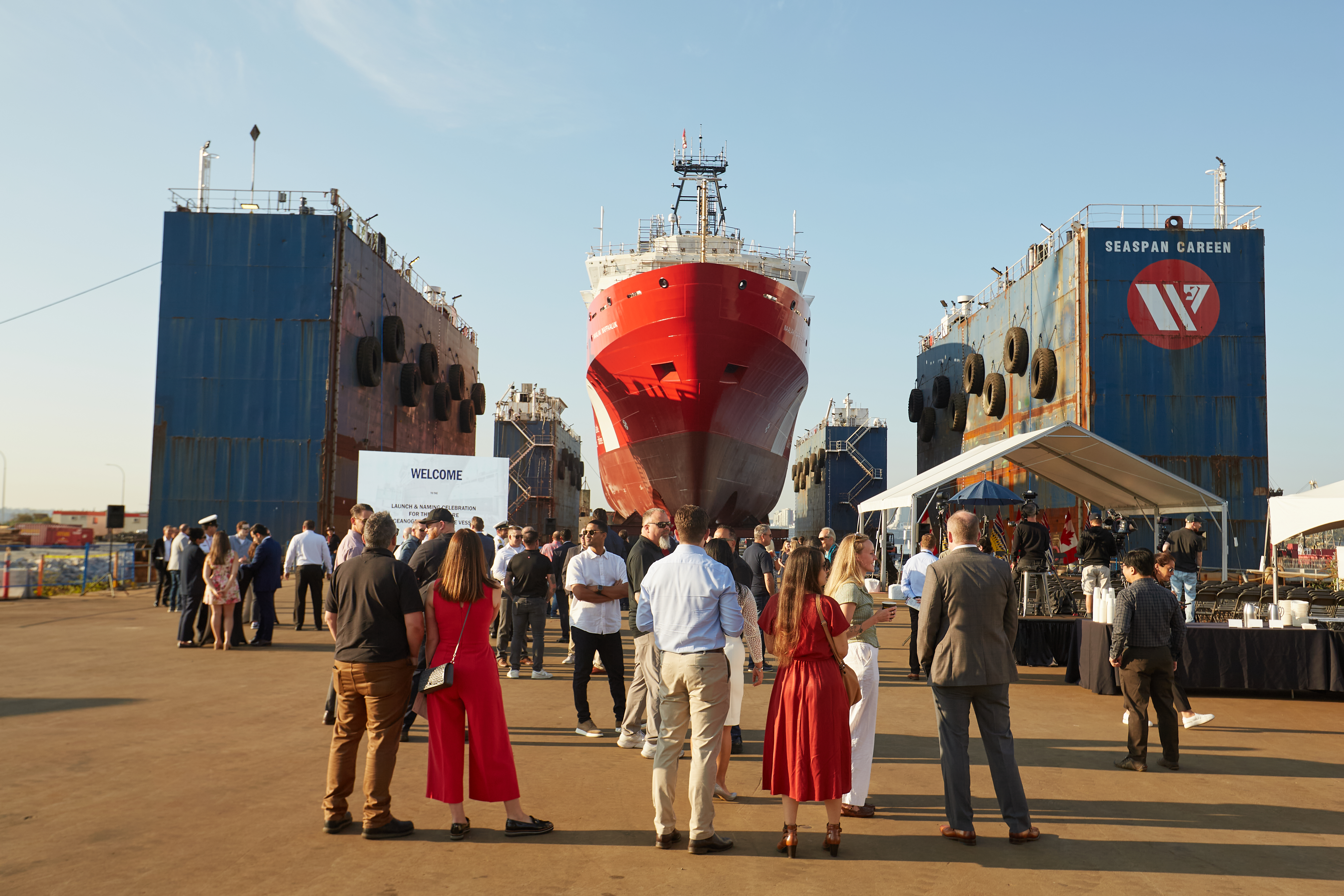Group of people standing on a dock in front of a large red and white ship positioned between 2 blue shipyard structures