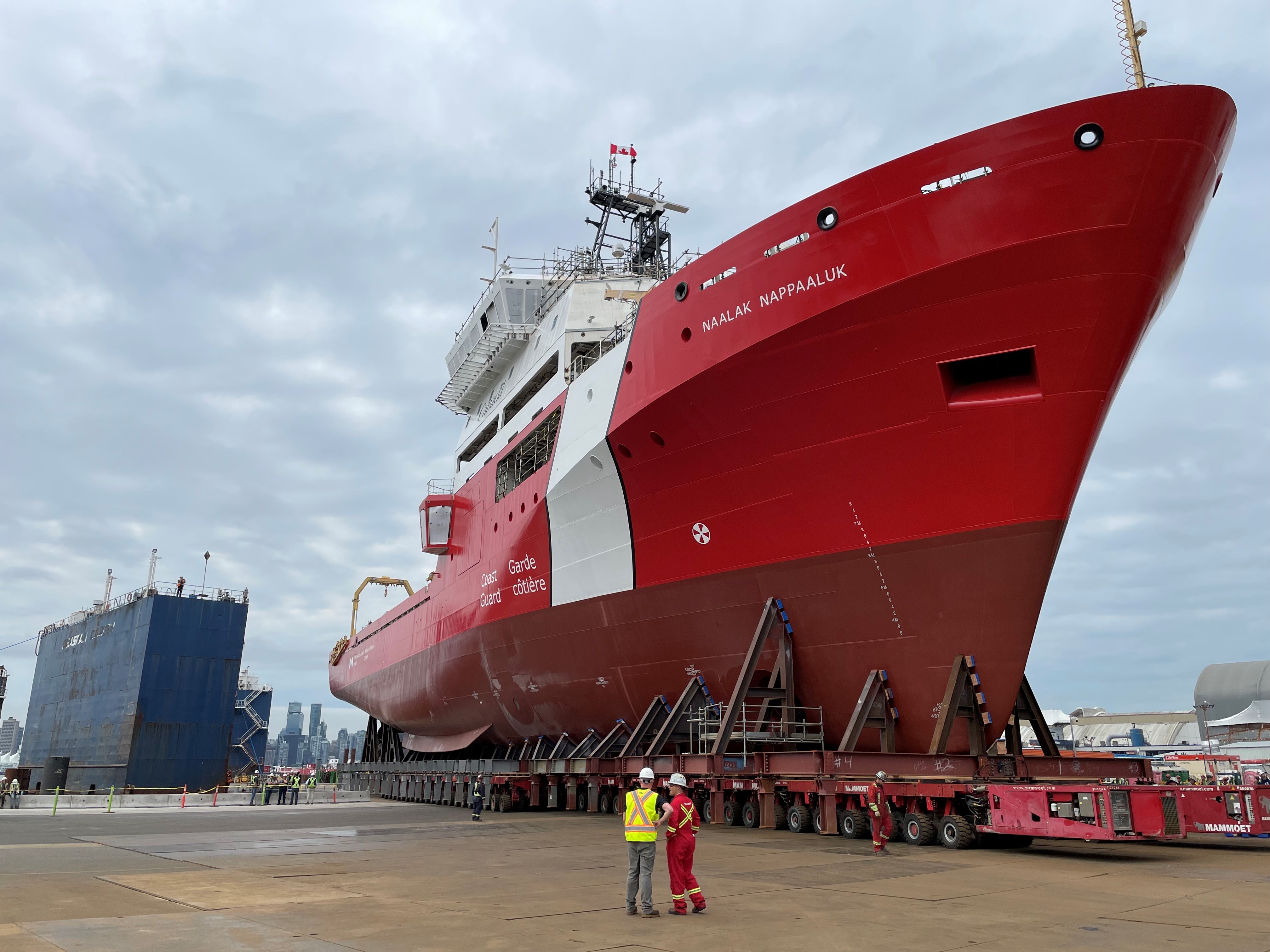 Large red and white ship named Naalak Nappaaluk on a dock, with 2 workers in safety gear standing in front and a blue shipyard structure in the background