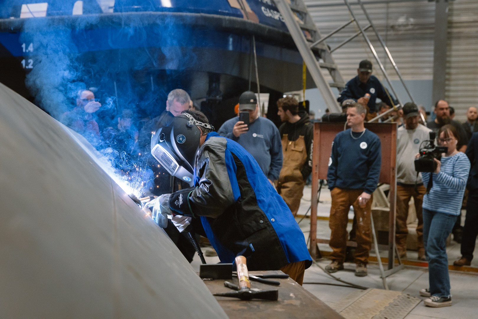Une personne portant un équipement de sécurité soude une surface métallique dans un atelier industriel, entourée d’observateurs.