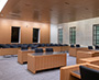 Enlarged image of the View from the hearing room dais showing counsel tables, audience seating, wood-panelled finishes, and large windows along a stone-clad wall.