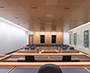 Enlarged image of the View from the hearing room dais showing counsel tables, audience seating, wood-panelled finishes, and large windows along a stone-clad wall.