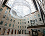 Enlarged image of the multi-storey atrium with stone walls, a glass skylight, a large suspended circular chandelier, and a curved staircase wrapping around a glass elevator leading to an upper-level walkway.