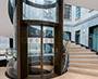 Enlarged image of the glass elevator surrounded by a curved staircase with glass railings, set within a multi-storey stone-clad atrium.