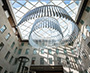 Enlarged image of the view upward through a multi-storey atrium with stone walls, a glass skylight, a large suspended circular chandelier, and a glass elevator beside the Supreme Court of Canada entrance.