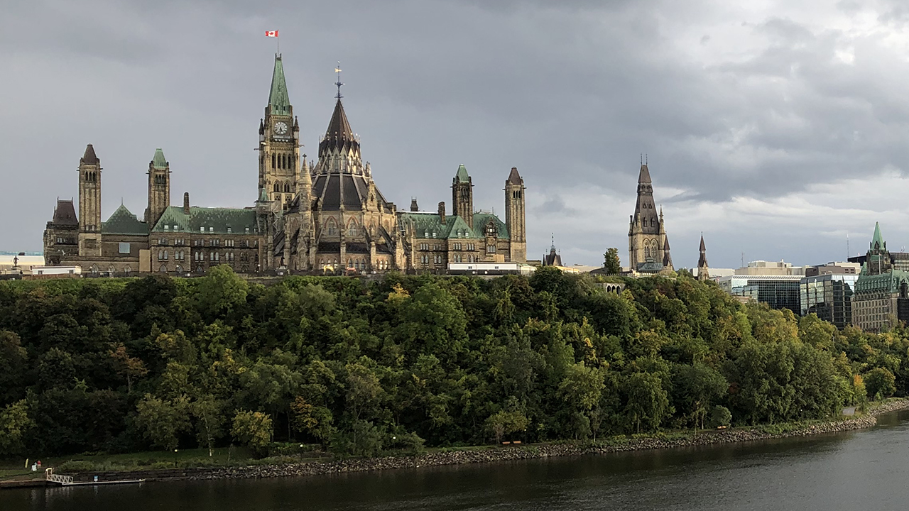 View of the Centre Block and its Peace Tower from the Ottawa Rriver. The Gothic building is atop a wooded escarpment on the bank of a river