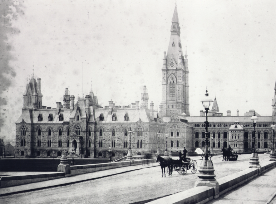 A historical photo of the completed West Block. An additional wing and tower have been added. Horses and carriages are on the road in front of the building