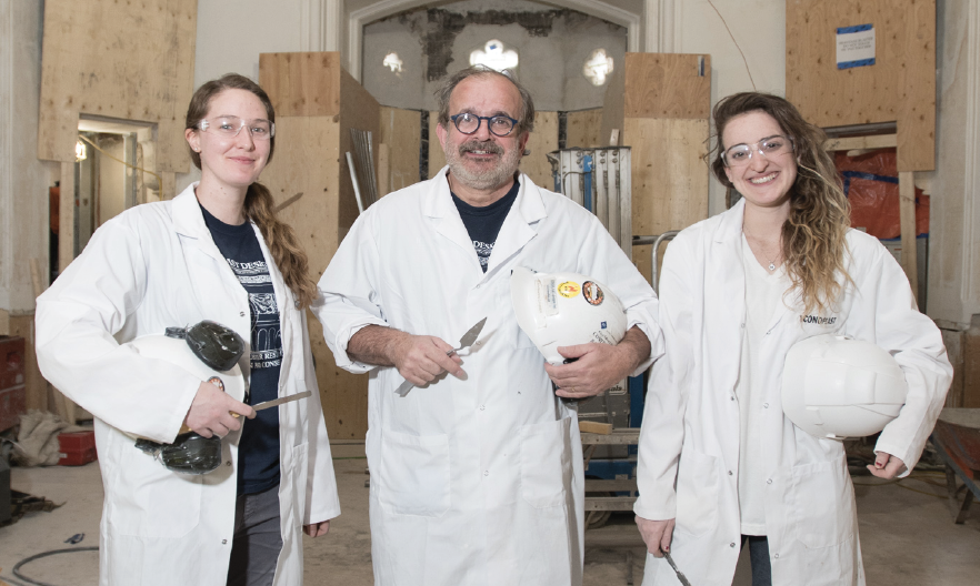 Un groupe de trois plâtriers debout côte à côte. Ils portent des manteaux blancs, des casques de protection et tiennent des outils pour manier le plâtre