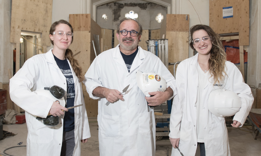 A group of three plaster tradespeople standing side by side. They are wearing white coats, hard hats, and holding plaster tools