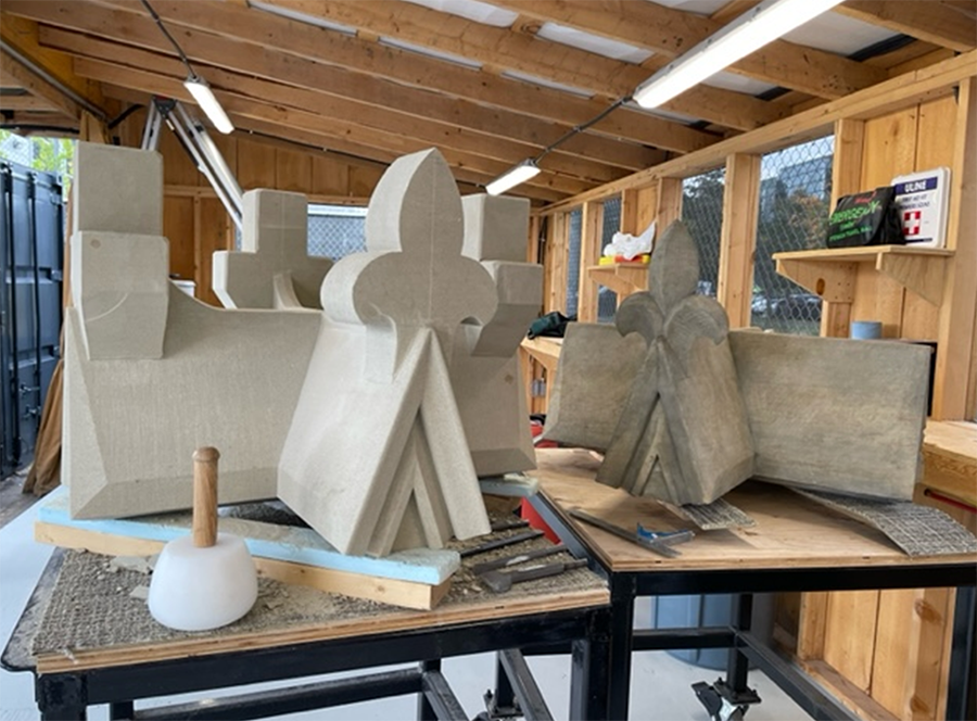 This is a side-by-side view of 2 cap stones. The stone on the right is the original, weathered and missing 3 of its 4 fleurs-de-lys. The stone on the left is a new replacement, partially carved, replicating the original design. Both stones rest on workbenches in a stonemasonry workshop