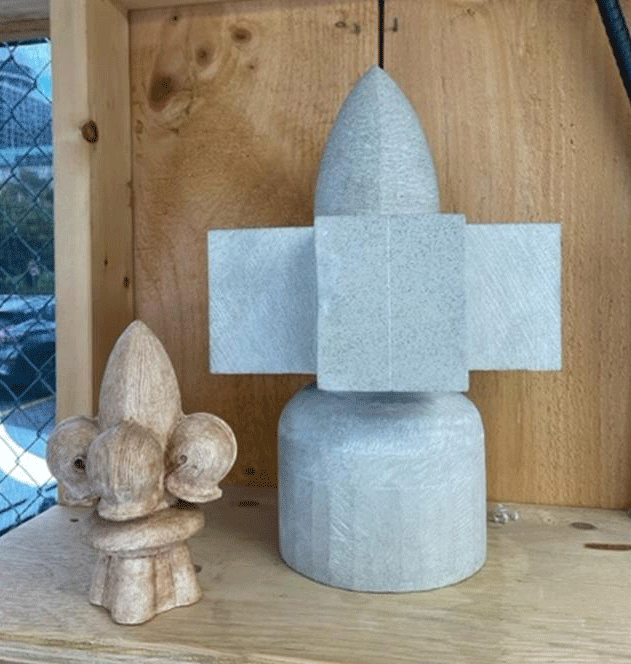 A partially carved replacement finial sits beside a smaller half-scale clay model in a workshop setting. The finial, shaped from limestone, will be installed on a decorative tower of the west elevation