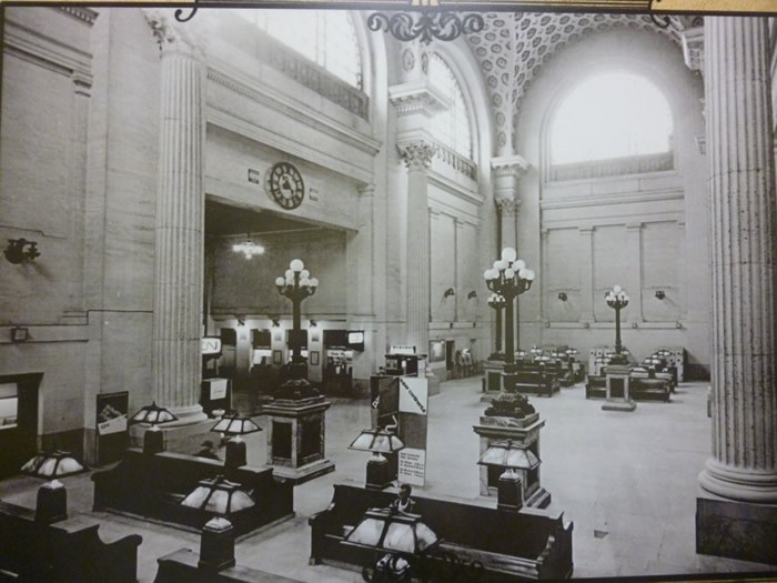 View enlarged image of a historical photo of a train station waiting area. People are sitting on benches.