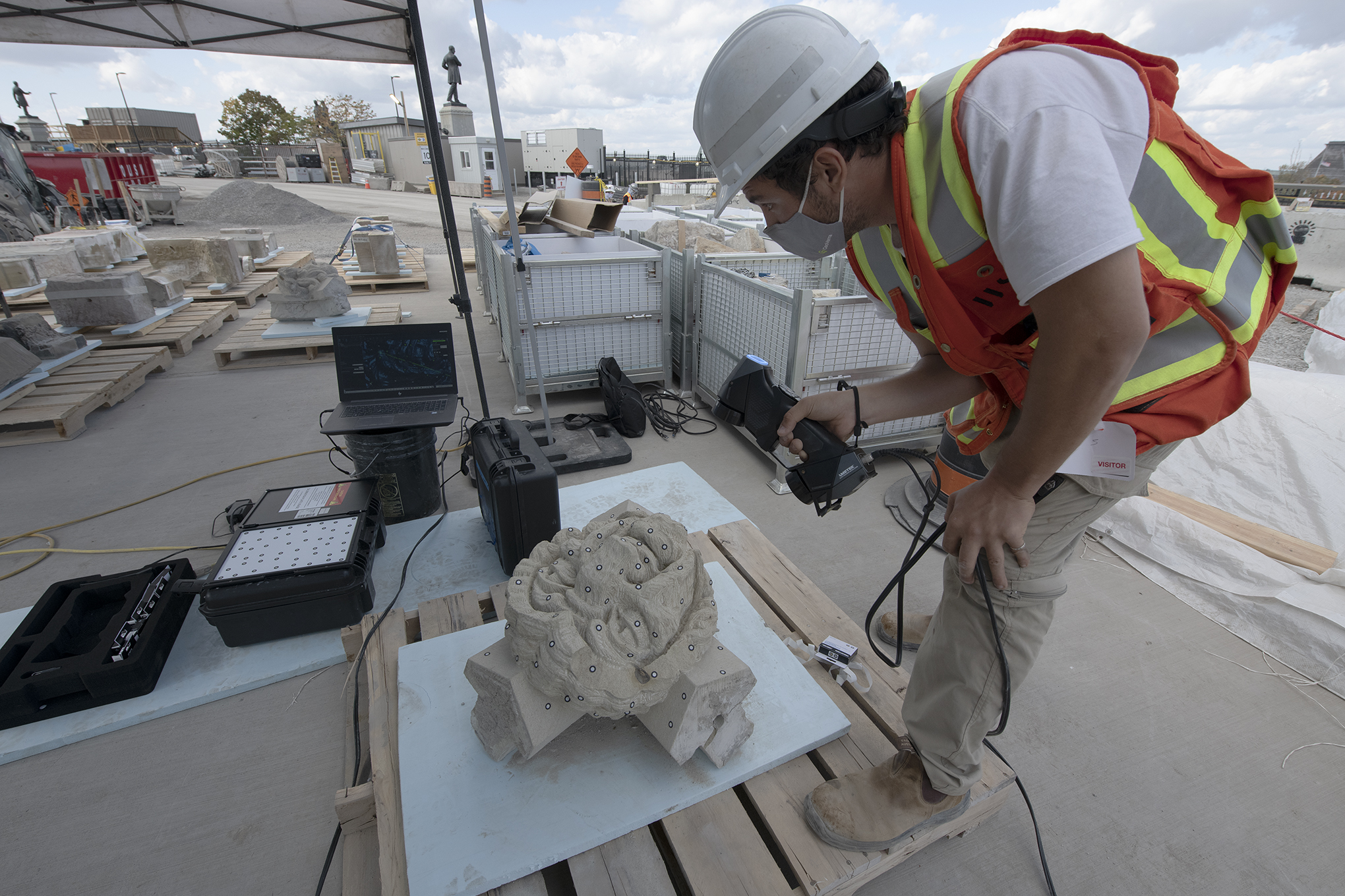 A man in a hardhat holds a 3D scanner over a stone carving. There are white dots on the stone carving.