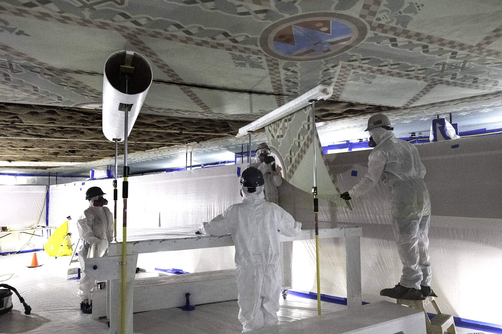 Three workers wearing protective equipment remove painted fabric from a ceiling and wind it onto a large spool.