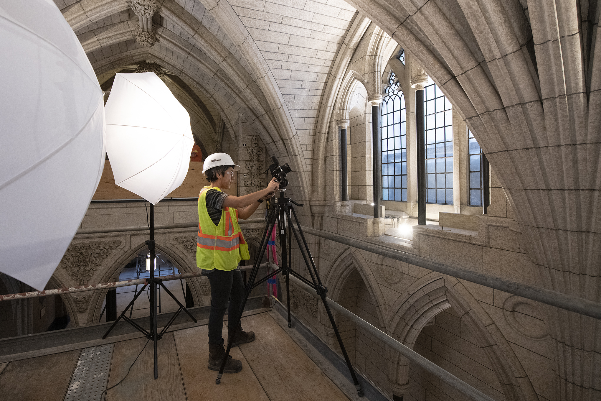 A worker is standing on scaffolding in an ornate room and taking photos of a carved vaulted stone ceiling.