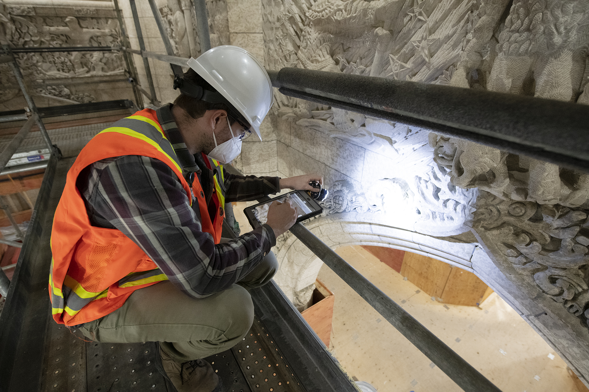 A man in a hardhat on scaffolding looks at carved stone and consults a tablet computer.
