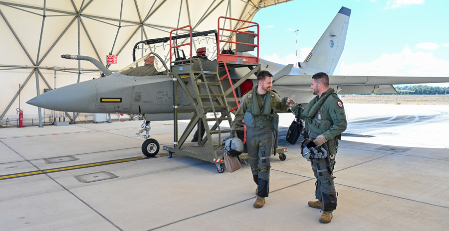 Deux pilotes portant une combinaison de vol verte descendent d’un avion gris à côté d’un escalier sous un hangar de toile.