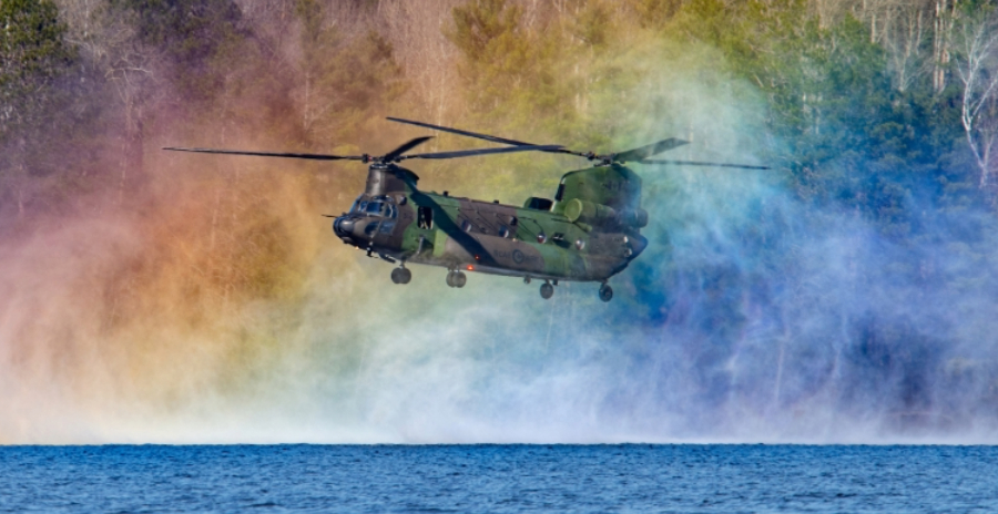 A CH-147F Chinook helicopter hovers low over a body of water, generating a spray with its rotors. A rainbow effect is visible in the mist.