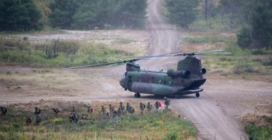 Canadian Armed Forces soldiers walk away from a CH-147F Chinook helicopter after disembarking on a dirt road in a forested training area.