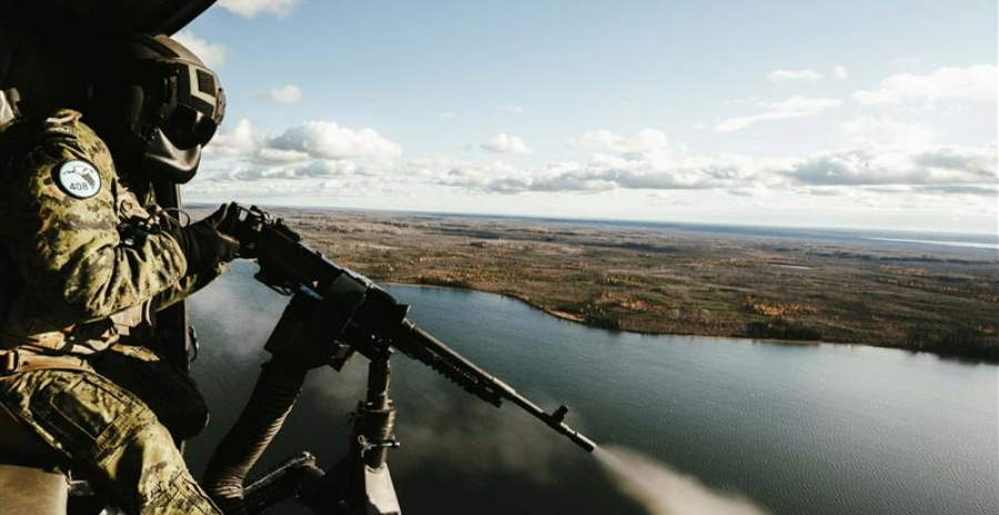 A door gunner soldier firing a mounted machine gun from the cabin of a CH-146 Griffon helicopter over a river and forested landscape.