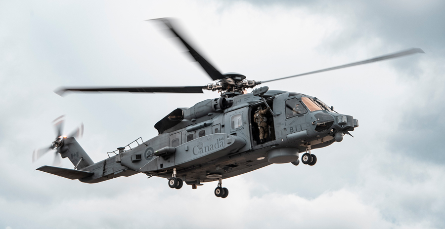 A grey military helicopter with its landing gear down hovers in the sky, with a crew member visible at the open side door.