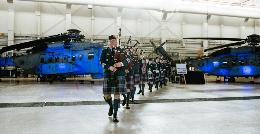 A military band in tartan kilts performs on bagpipes and drums inside a hangar, with helicopters and event signage in the background.