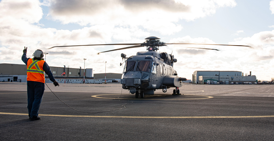 A person wearing a safety vest and helmet directs a helicopter preparing for takeoff on a wide tarmac under a cloudy sky.