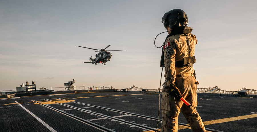 A uniformed crew member stands on a naval vessel’s flight deck guiding a helicopter hovering nearby.