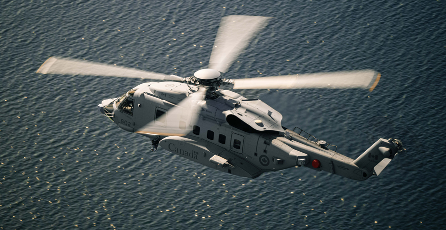 A Canadian military helicopter flies over open water, its rotor blades blurred with motion in the sunlight. 