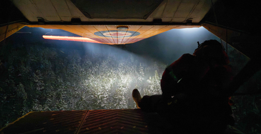 A person sits at the open ramp of an aircraft flying over a forest at night. The aircraft’s lights illuminate the snowy trees below.