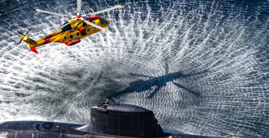 A yellow and red search and rescue helicopter hovers above the conning tower of a surfaced submarine and a person stands atop the submarine.